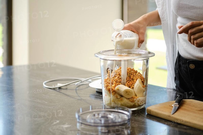 Ingredients are being added to a blender for smoothies. A person is pouring milk into the container filled with ripe bananas and sea buckthorn