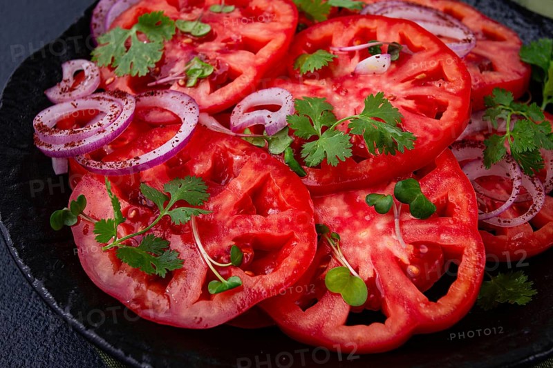 Freshly sliced tomatoes and red onions, garnished with herbs, on a black plate, summer salad, no people