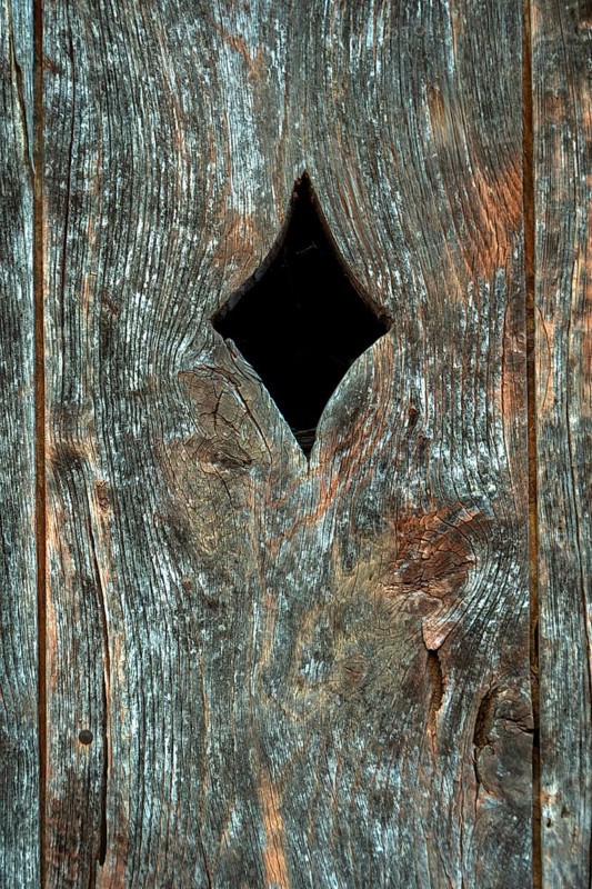 A close-up view of a weathered wooden panel, featuring a diamond-shaped opening cut into the surface. The wood displays signs of age with a rich texture and grain