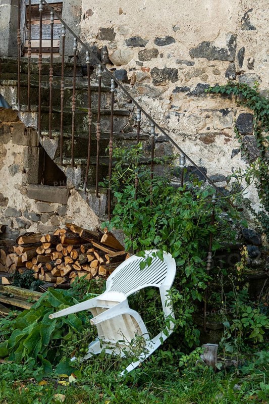An unkempt outdoor area features a white chair leaning against a weathered stone wall, surrounded by creeping vines and overgrown plants. Nearby, a stack of firewood illustrates neglect. France