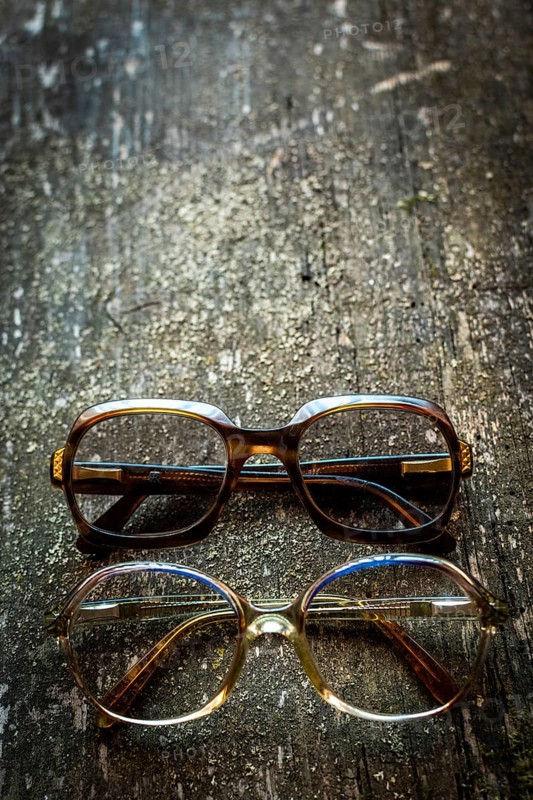 A collection of vintage eyeglasses rests on a rustic wooden surface, illuminated by warm lighting that highlights their unique designs and charm, evoking a sense of nostalgia