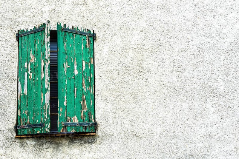 Weathered green shutters stand slightly ajar on a textured white wall, showcasing the charm of an old building and reflecting years of history and character