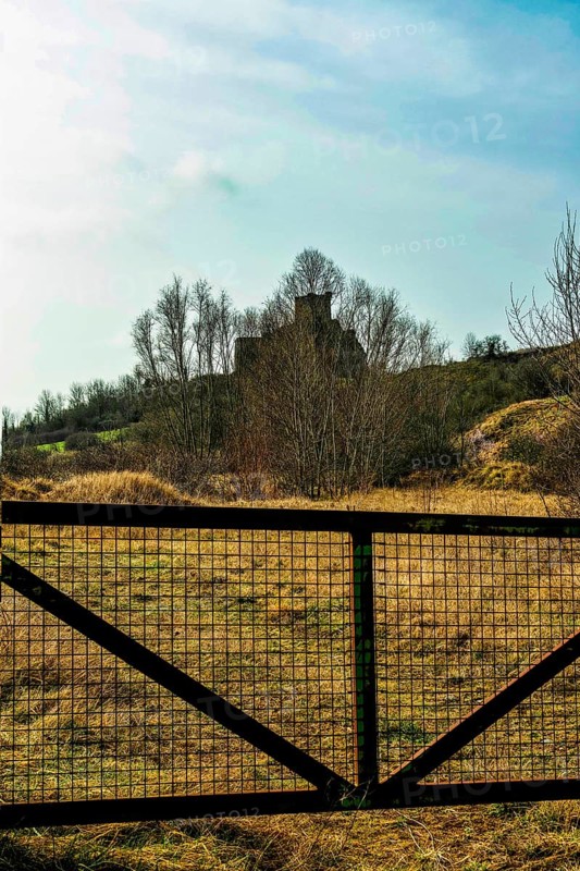 A picturesque view captures the ruins of a castle atop a hill, framed by a metal gate. The landscape features dry grass and scattered trees, France