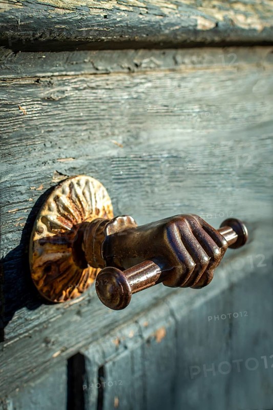 A distinctive door handle resembling a hand grips a rustic wooden door, featuring intricate details and a charming patina. The warm light enhances the texture and design