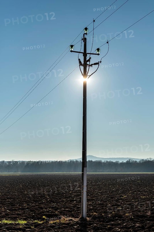 A power pole stands tall in the foreground, with sunlight shining directly behind it, creating a glowing silhouette. The background features an expansive field under a blue sky. France