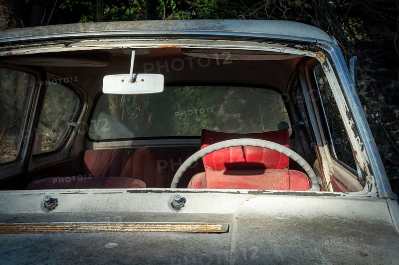 A deserted vintage car sits in a state of disrepair, showcasing a cracked windshield and a faded interior. The red seat adds a pop of color under soft, natural light