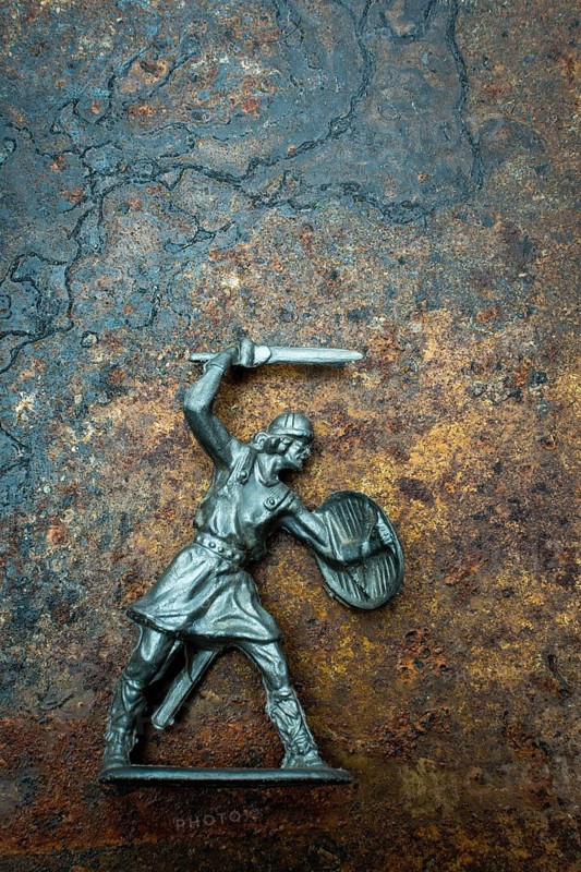 A detailed warrior figure stands ready for battle, gripping a sword in one hand and a shield in the other. Its metallic texture contrasts with the earthy backdrop, suggesting a fierce engagement