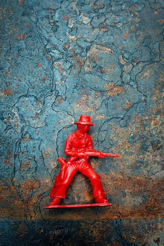 A small red plastic toy cowboy stands firmly on a rustic blue and brown textured background. The figure is equipped with a weapon