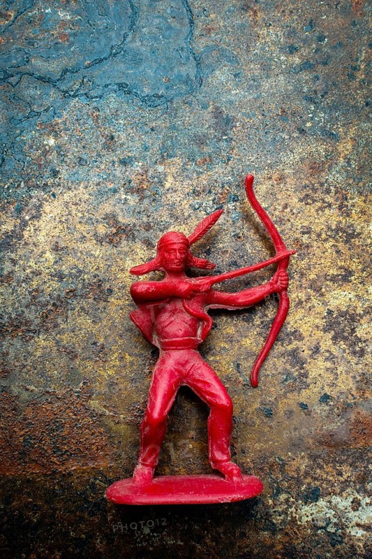 A red plastic toy archer represents a Native American figure, posed with a bow and arrow on a textured, rust-colored surface. T