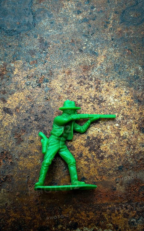 A small green plastic toy cowboy stands firmly on a rustic blue and brown textured background. The figure is equipped with a weapon