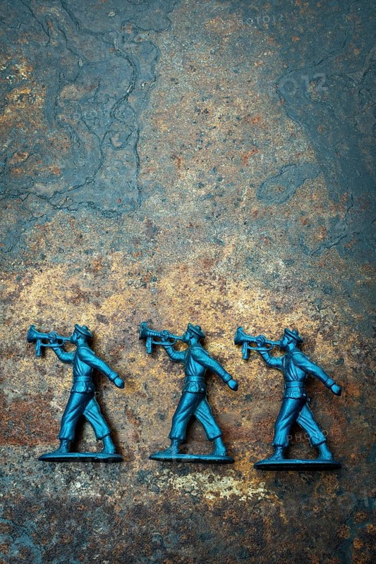 Three small blue toy soldiers are positioned on a rough, textured surface, representing scenes from childhood games. Their sturdy posture and details trigger nostalgic memories of playtime adventures