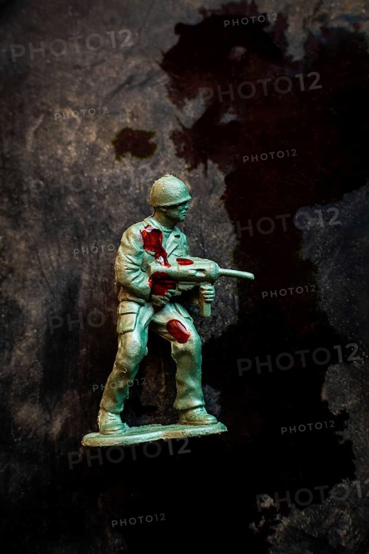 A green soldier figurine holds an assault weapon while displaying battle wounds marked by red stains. The dark backdrop serves to enhance the gravity of war and its emotional toll