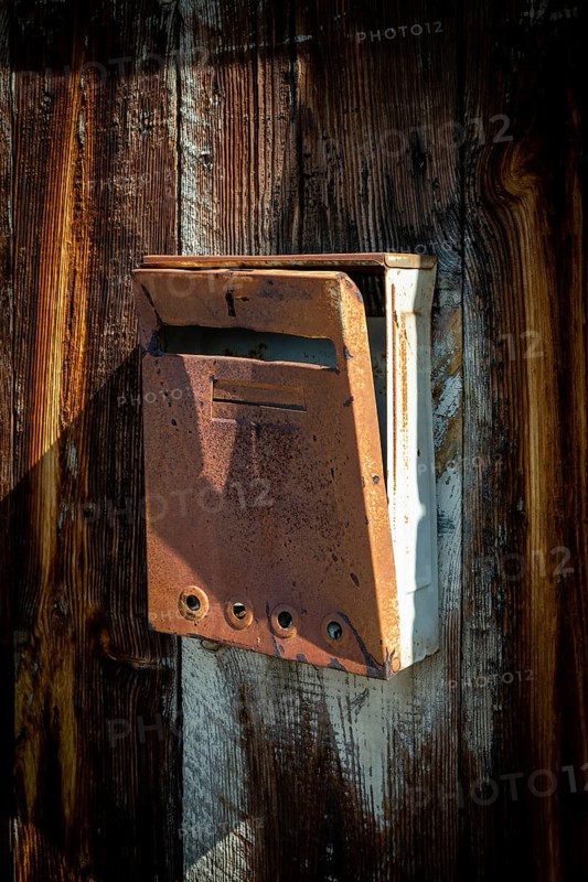 A vintage mailbox is mounted on a weathered wooden fence, bathed in warm sunlight. Its rusted surface and unique design tell a story of history and age, attracting attention. France