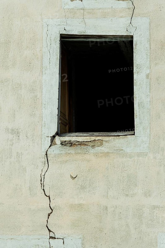 A cracked wall features an empty window opening, creating a stark contrast against the faded texture of the surrounding surface in a dimly lit environment