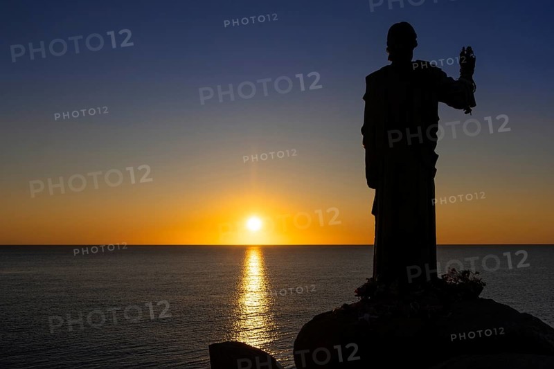 The sun rises on the Costa Rei, a stretch of coastline on the Italian Mediterranean island of Sardinia. In front of it is the silhouette of a 1.80 metre high bronze statue of Christ called the Blessing Christ of Santa Giusta, also known as Christ the Navigator, Monte Nai, Costa Rei, Sardinia, Italy