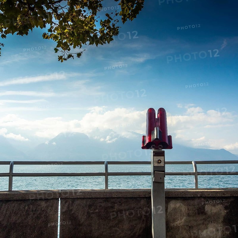 A telescope stands on the lakeside walkway in Vevey, Switzerland, overlooking Lake Genova. The clear sky and distant mountains create a picturesque backdrop for visitors. Switezerland