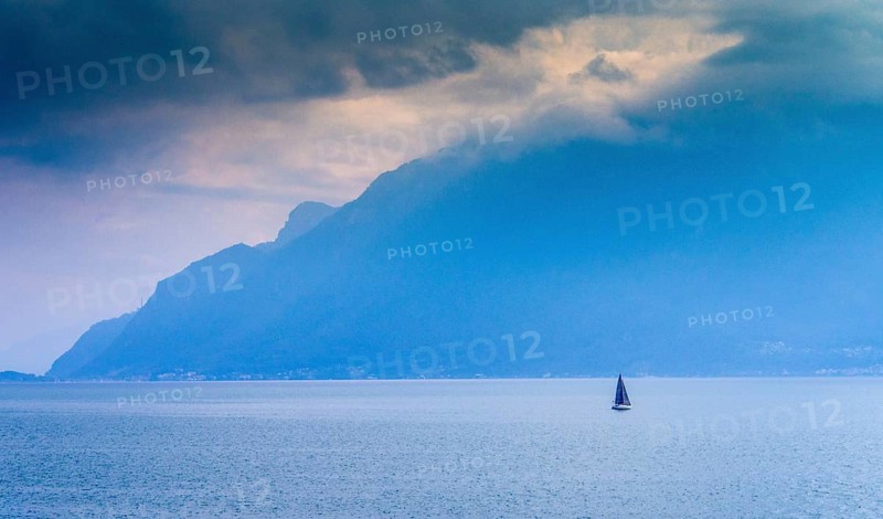 A lone sailboat navigates the tranquil waters of Lake Geneva surrounded by shadowy mountains. The atmosphere is calm yet dramatic under the cloudy sky. Switzerland