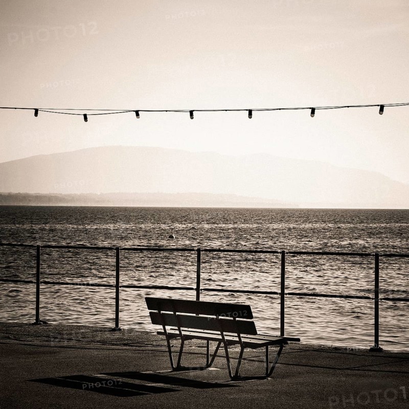 A solitary bench sits by the lakeside promenade in Nyon, Switzerland. Gentle waves ripple in Geneva Lake while distant mountains create a tranquil backdrop, enhancing the peaceful atmosphere