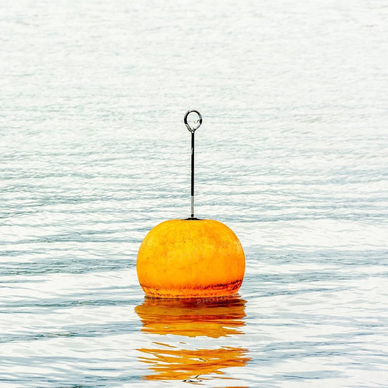 A bright orange buoy stands upright on the tranquil waters of Lake Geneva in Switzerland, surrounded by stunning natural scenery. Soft ripples reflect the buoy's color as it peacefully floats