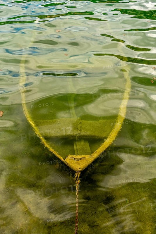 A small boat partially submerged in a calm lake in France showcases its weathered hull beneath the water's surface, surrounded by tranquil reflections and natural beauty