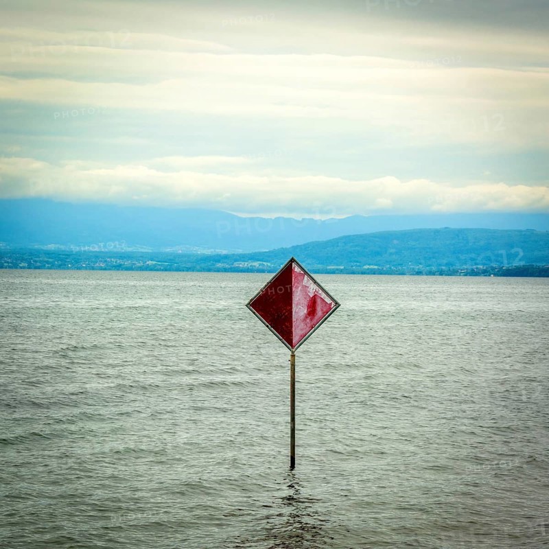 A prominent red information sign rises above the water of Lake Geneva, indicating safety or navigation. The scenery features a serene lake with distant hills visible against a cloudy sky. Switzerland