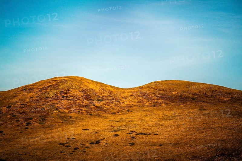 Two gentle hills rise from a golden landscape under a bright blue sky. Wispy clouds streak across the sky, adding depth to the peaceful scene. The setting radiates tranquility. Puy de Dome. Auvergne Rhone Alpes. France