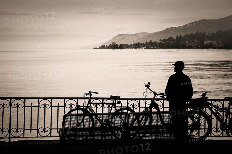 A tourist stands at the edge of Lake Geneva, captivated by the serene waters and distant hills. Bicycles are parked nearby, creating a peaceful atmosphere for exploration at sunrise. Switzerland