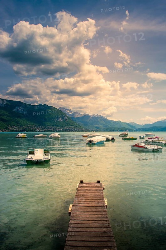 A wooden pontoon extends over the tranquil waters of Annecy Lake, surrounded by mountains and dotted with colorful boats. The sky is partially covered by clouds, creating a serene atmosphere. Haute Savoie. Auvergne Rhone Alpes. France