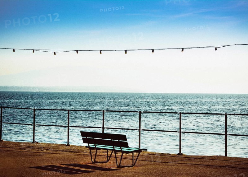 A bench is positioned along the promenade beside Geneva Lake in Nyon, Switzerland, providing a peaceful spot to enjoy stunning views of the calm water and clear blue sky