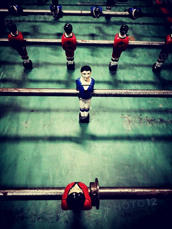 A lively table football game features diverse player figures in red, blue, and black, positioned on a green playing surface. Players compete for victory in a friendly match
