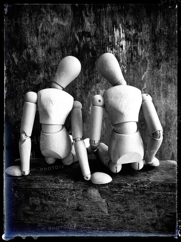 Two wooden figures are positioned closely together, facing away from the viewer. They sit on a rustic wooden surface, showcasing their joint form in a serene and artistic manner