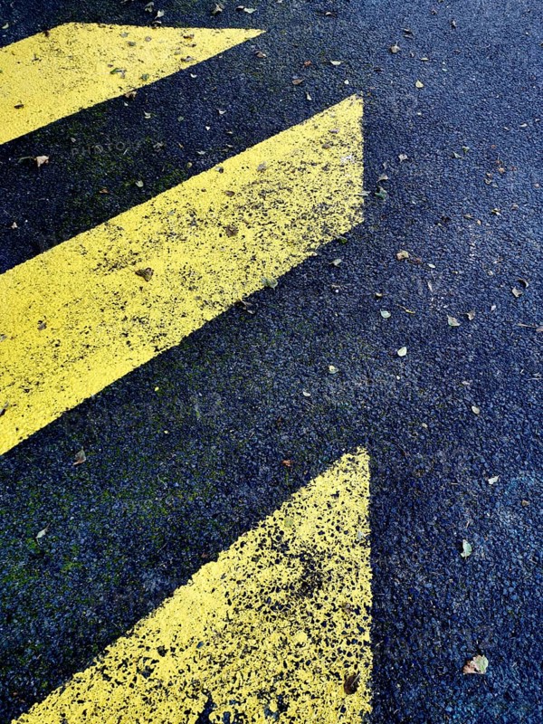 Yellow directional markings stretch across a dark asphalt surface, creating a striking contrast. Scattered autumn leaves add a natural element to the scene, enhancing its texture and depth