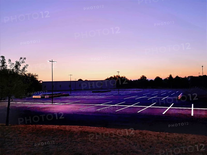 A quiet parking lot in France at dusk, illuminated by streetlights. The clear sky transitions from deep blue to vibrant orange, casting an inviting glow over the empty spaces. France