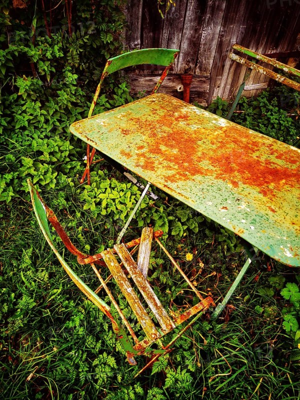 A rusty green table and a broken chair lay forgotten in thick, overgrown grass, evoking a sense of nostalgia and nature reclaiming human artifacts in a quiet outdoor area