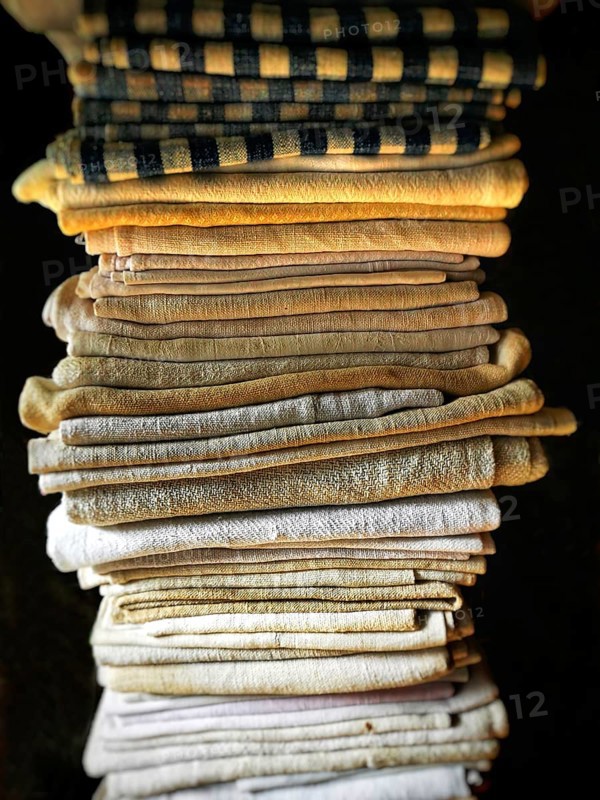 A neat stack of folded textiles in various shades of beige, cream, and a hint of black is illuminated by soft, natural light, enhancing their textures and warmth