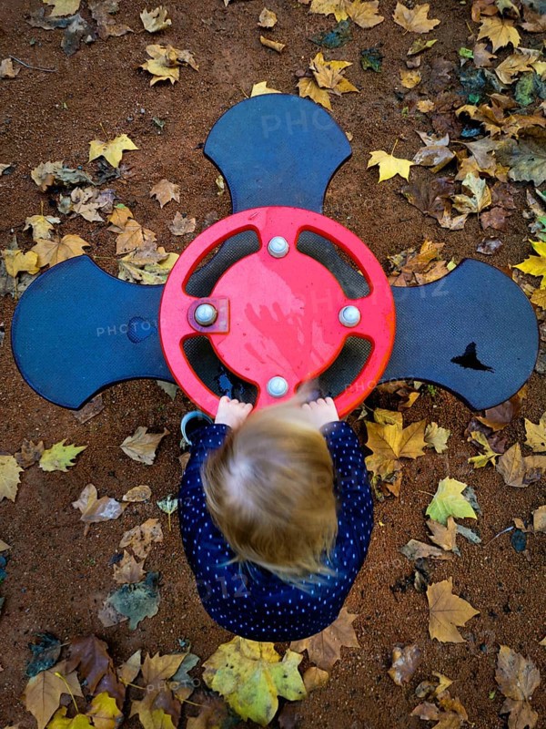 A young child is playing with a vibrant red spinner at a playground, surrounded by fallen yellow and brown leaves, capturing the essence of autumn joy and exploration