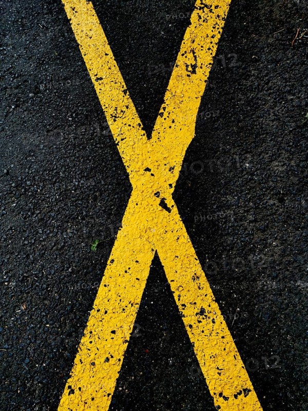 A pattern of yellow crossing lines painted on a dark asphalt surface emphasizes a bold visual contrast. The geometric marks suggest direction and movement on a city road