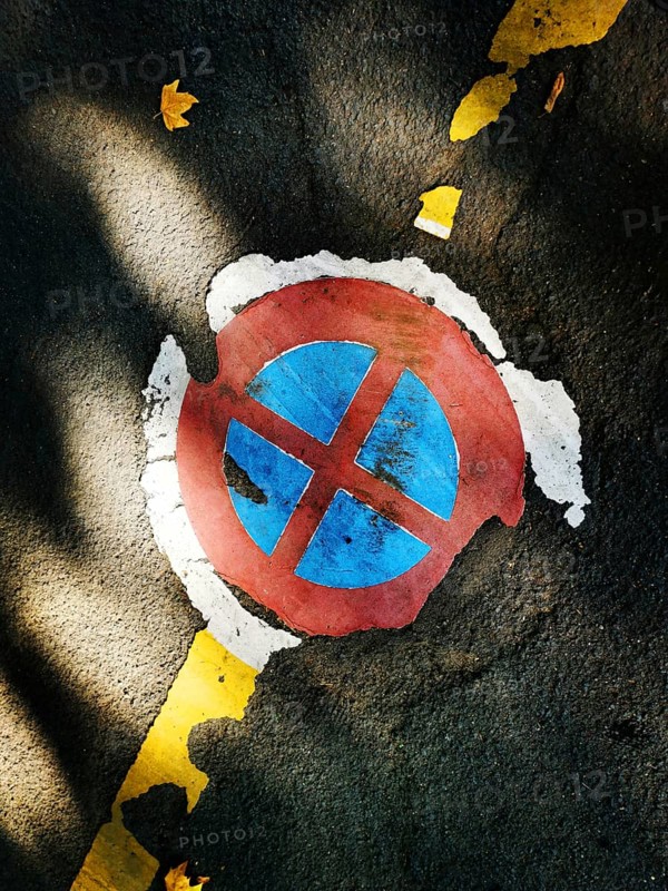 A circular parking symbol with red and blue colors appears worn and cracked on an asphalt surface. Yellow lines border the sign, and fallen leaves add a touch of autumn
