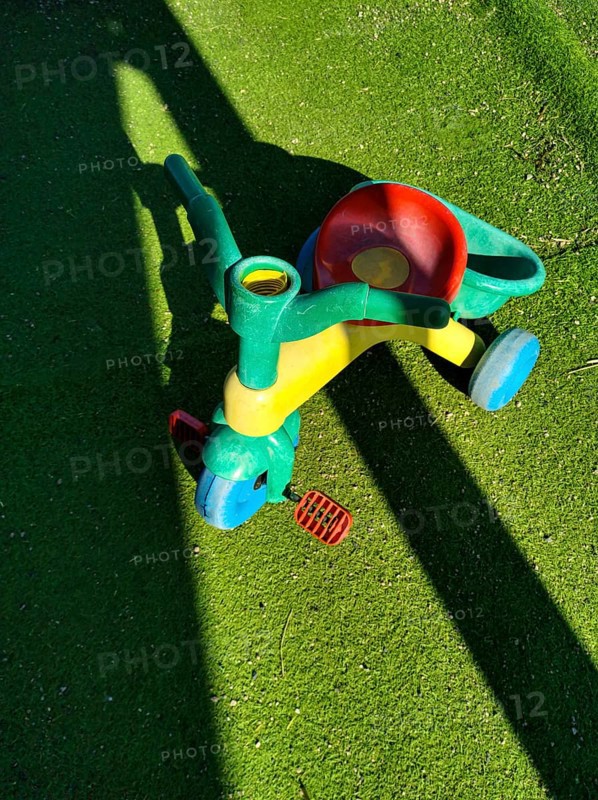 A vibrant, multi-colored tricycle sits on lush, green artificial grass, casting a shadow in bright sunlight. Its playful design invites children's imagination and outdoor fun