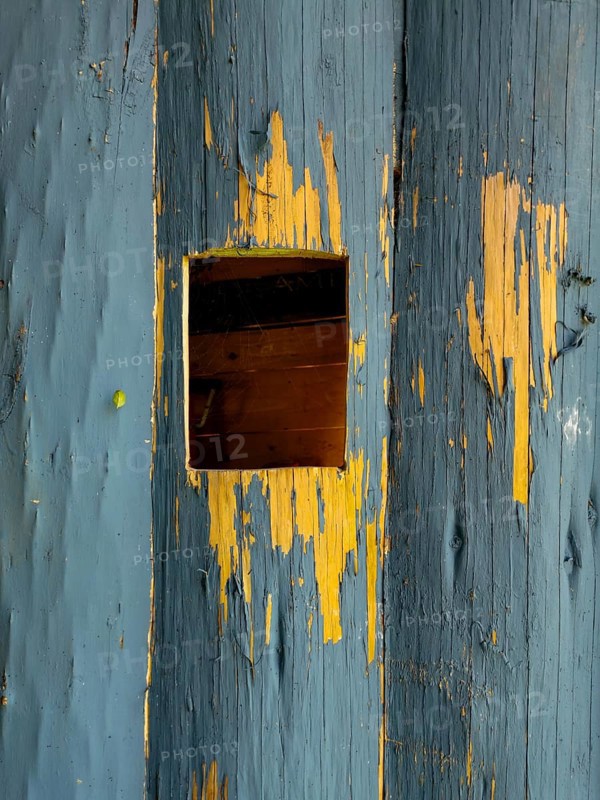 A small square opening in a weathered blue wooden surface showcases the contrast of peeling paint and exposed wood grains. The texture tells a story of age and exposure