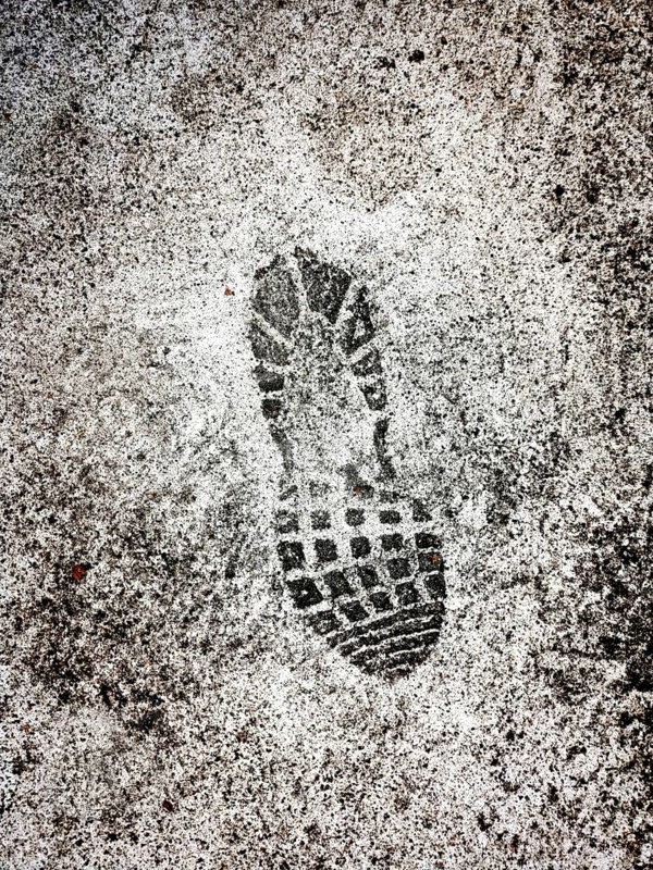 A distinct shoe print is marked on a wet surface, showing details of the tread pattern. Surrounding areas are blurred, hinting at a bustling urban environment after rain