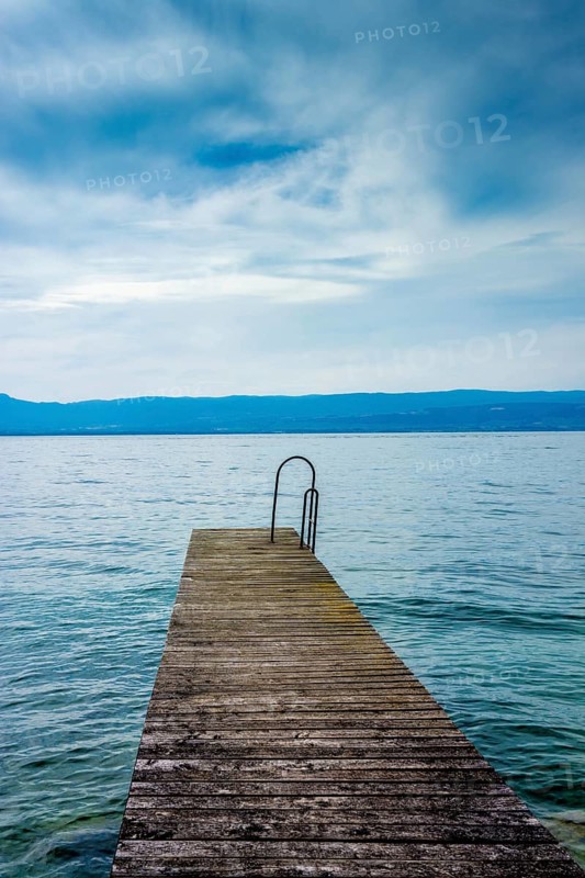 A sturdy jetty extends into the serene waters of Lake Geneva, surrounded by mesmerizing blue hues and a tranquil atmosphere. Perfectly captures the beauty of Haute-Savoie on a peaceful day, Auvergne Rhone Alpes, France