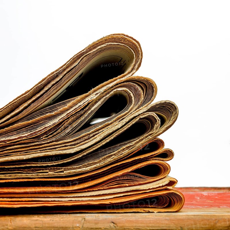 A stack of newspapers lies neatly on a white background, displaying an assortment of folded pages with printed articles and images. The newspapers show signs of wear and age, indicating their history
