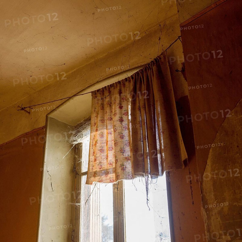 Sunlight streams through a dusty window adorned with worn curtains, highlighting the cobwebs in a neglected room. Dust particles float in the air, evoking a sense of nostalgia