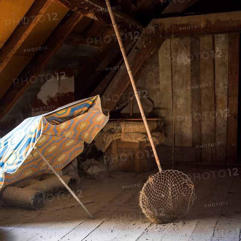 Sunlight filters through an attic, illuminating a colorful sunshade and a butterfly net alongside dust-covered wooden beams, creating a tranquil, nostalgic atmosphere