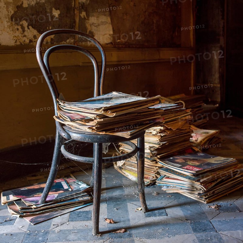 An old chair is perched beside a towering stack of newspapers, both covered in dust, in a dimly lit room that suggests neglect and the passage of time, evoking memories of a bygone era