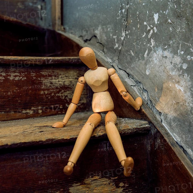 A wooden mannequin is seated casually on a weathered staircase, surrounded by rustic decor and peeling plaster. The scene captures a blend of artistry and nostalgia in a unique interior space