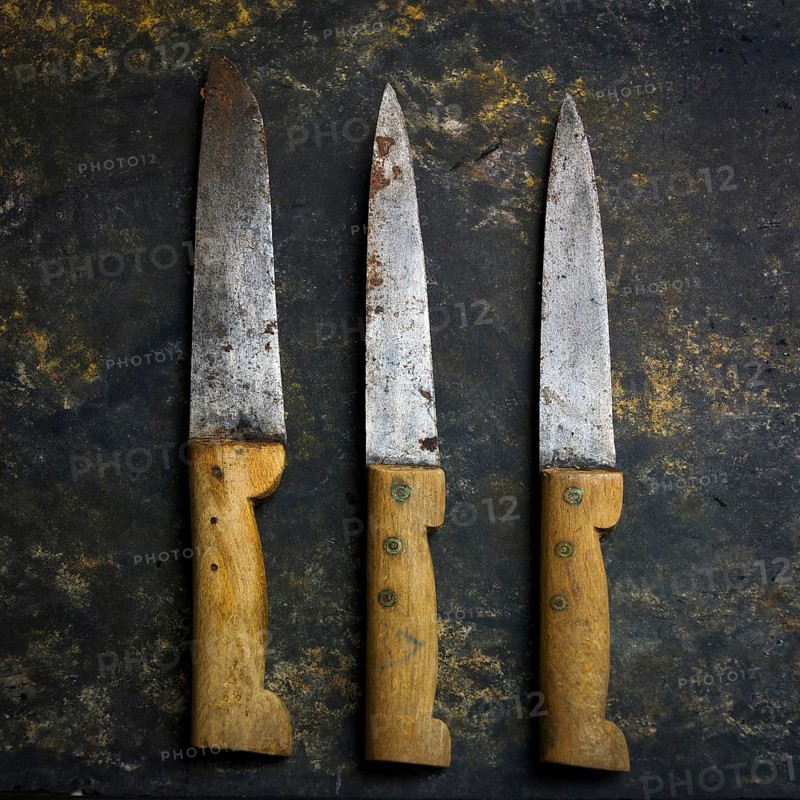 Three vintage kitchen knives lie on a dark, textured surface, their worn blades reflecting use, showcasing the craftsmanship of kitchen tools and the passage of time