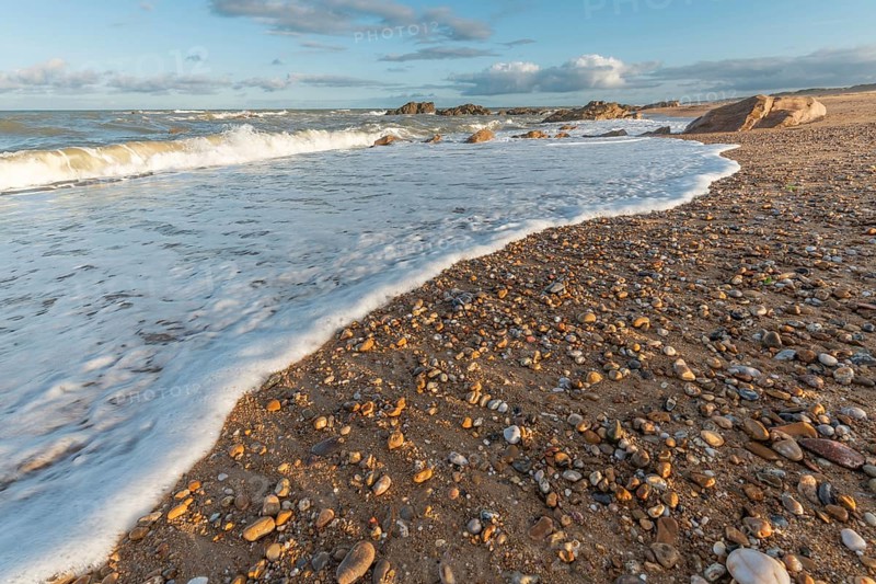 A wide sandy beach stretches out with scattered pebbles and beach shells. Gentle waves crash against the shore under a partially cloudy sky during late afternoon. Plage de l'Aubraie, Les Sables d'Olonne, Vendee, France