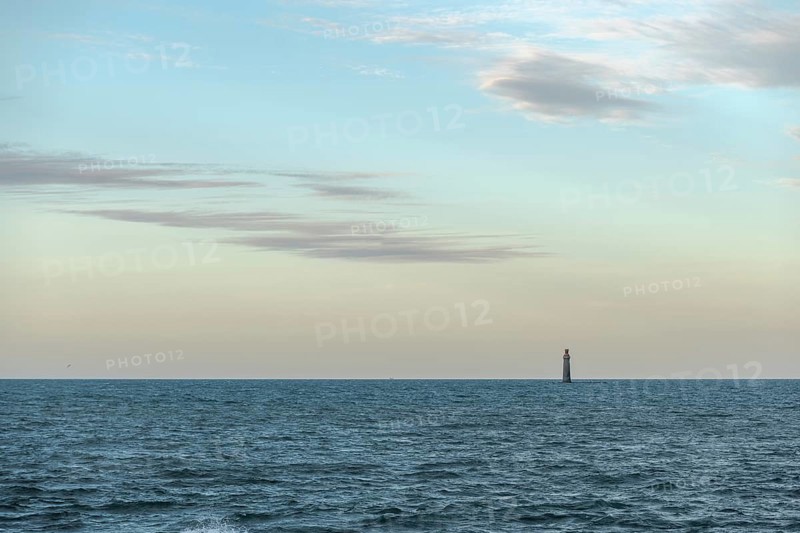 A lighthouse rises from the gentle waves of the ocean, surrounded by a tranquil seascape. Soft clouds drift over the sunset, creating a peaceful atmosphere. Les Sables d'Olonne, Vendee, France